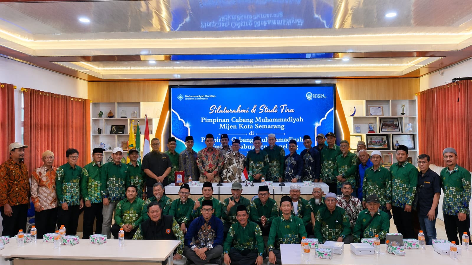 Rombongan pengurus PCM Mijen Semarang berseragam batik hijau berfoto bersama jajaran PCM Muntilan dengan latar layar bertuliskan Silaturahmi dan Studi Tiru dalam rangkaian napak tilas sejarah Muhammadiyah.