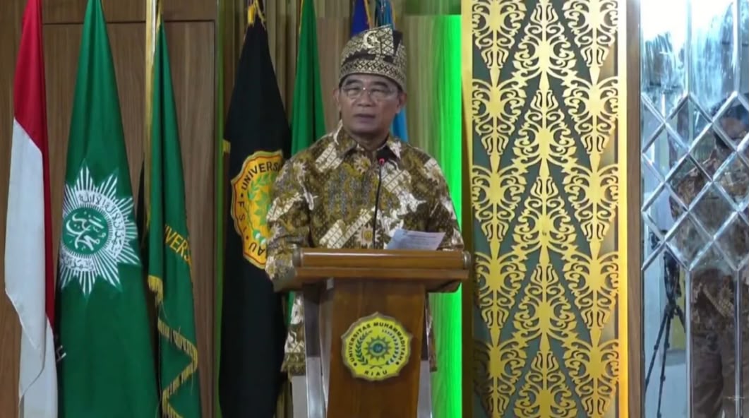 Muhadjir Effendy memberikan sambutan terkait program pemerataan kesehatan Muhammadiyah di podium Universitas Muhammadiyah Riau (UMRI). Pria tersebut mengenakan batik cokelat dan penutup kepala tradisional (tanjak) emas, berdiri di depan bendera Merah Putih serta bendera organisasi.