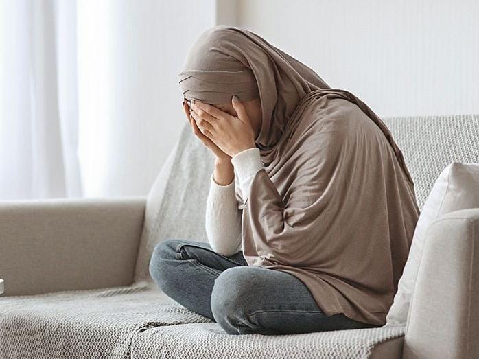 Sesosok wanita berhijab cokelat muda duduk bersila di sofa kain sambil menutupi wajahnya dengan kedua telapak tangan, mengilustrasikan kondisi stres kerja yang membutuhkan kemampuan untuk kelola emosi dan hati agar meraih ketenangan jiwa.