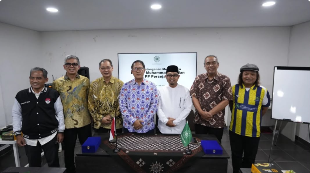 Jajaran pengurus LPO PP Muhammadiyah dan Persejasi berfoto bersama setelah penandatanganan MoU program gaya hidup sehat muhammadiyah lewat sepak bola berjalan kaki. Tujuh orang pria berdiri di depan layar digital di Ruang Aula Kantor PP Muhammadiyah Yogyakarta, dengan meja berbalut kain motif matahari dan bendera kecil di depannya.