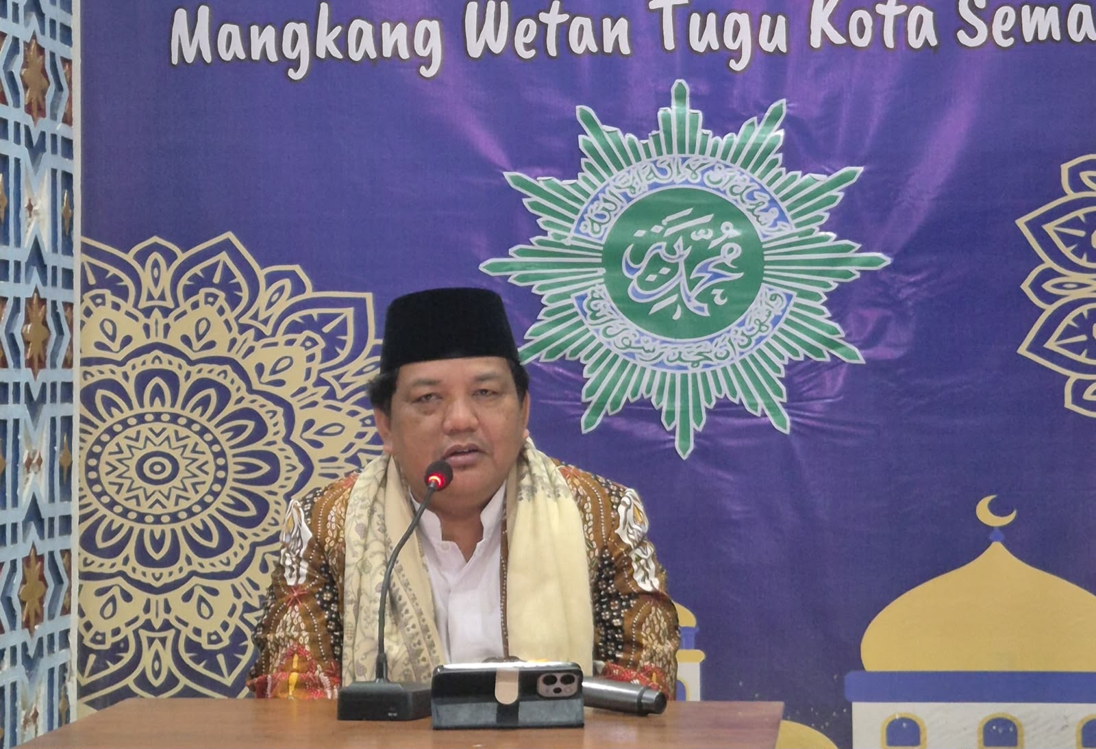 Dr. H. AM Jumai, S.E., M.M. duduk menyampaikan ceramah mengenai kunci hidup tenang di depan sebuah mikrofon meja. Tokoh tersebut mengenakan kemeja batik, sorban krem di leher, dan peci hitam dengan latar belakang spanduk ungu bertuliskan "Mangkang Wetan Tugu Kota Semarang" serta logo matahari Muhammadiyah hijau-putih.