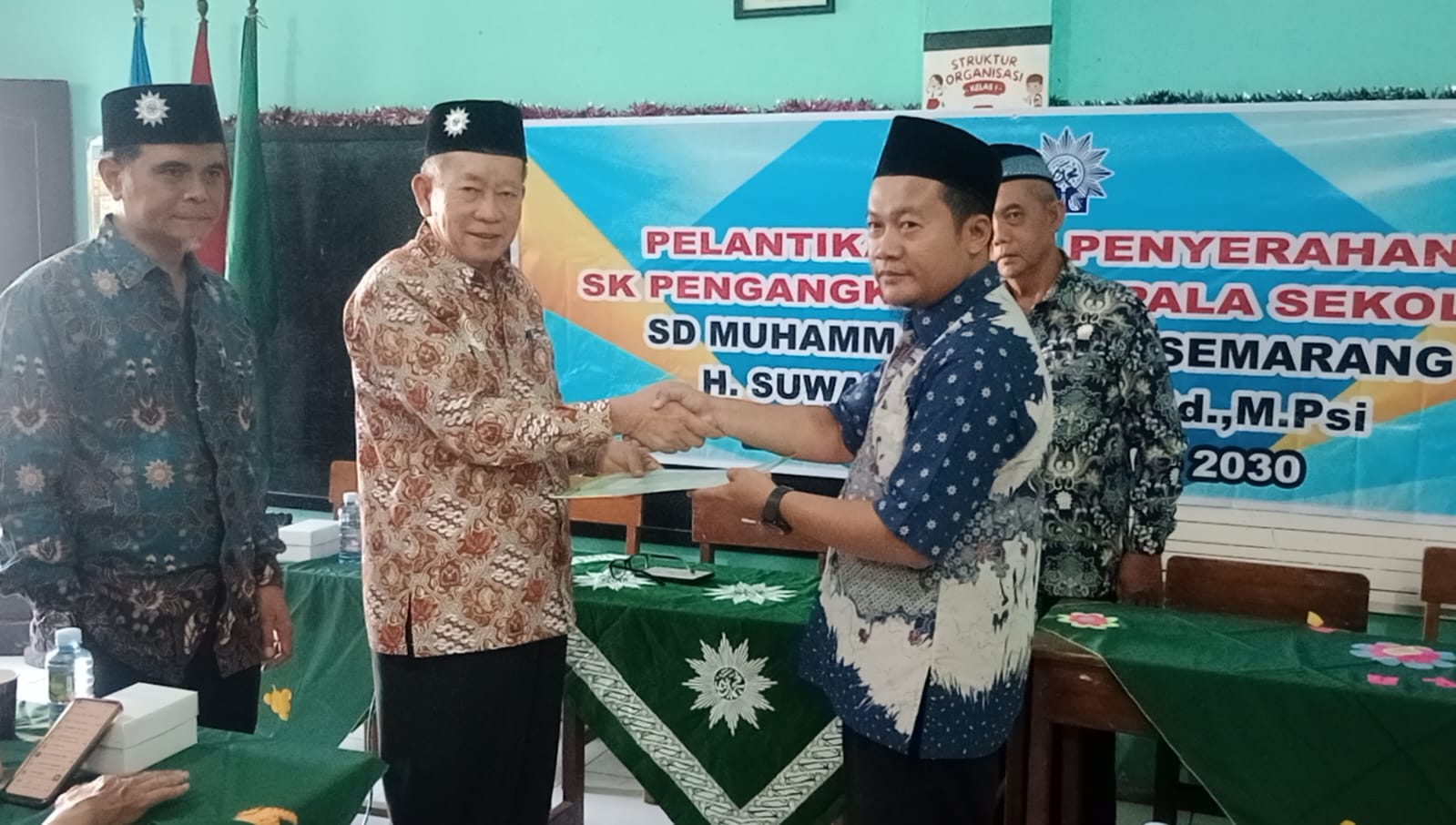 Prosesi jabat tangan antara pimpinan organisasi dan kepala sekolah baru saat penyerahan SK pelantikan dengan misi mencetak generasi qurani di SD Muhammadiyah 10 Semarang. Empat orang pria mengenakan batik dan peci berdiri di depan spanduk acara bernuansa biru-kuning.