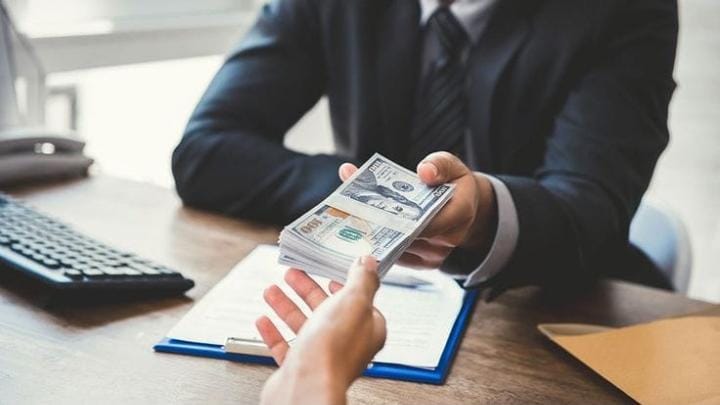Foto close-up tangan seorang pria bersetelan jas formal yang menyerahkan seikat tebal uang kertas pecahan 100 Dolar AS kepada orang lain di atas meja kerja. Gambar ini merepresentasikan ilustrasi transaksi pinjaman tidak sehat maupun praktik suap yang memicu dampak buruk riba bagi tatanan sosial ekonomi.