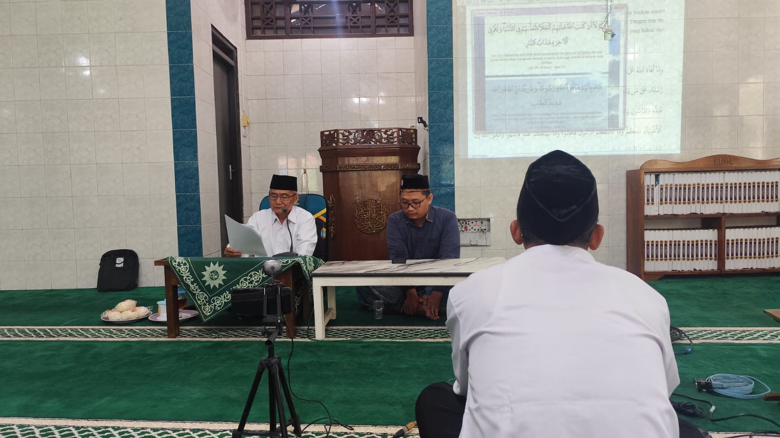 Ust. Dr. H. Zuhad Masduki, M.A. menyampaikan materi kajian di depan mimbar masjid dengan fasilitasi layar proyektor yang menampilkan ayat Al-Qur'an, memberikan pemahaman mendalam tentang pelajaran bani nadhir.