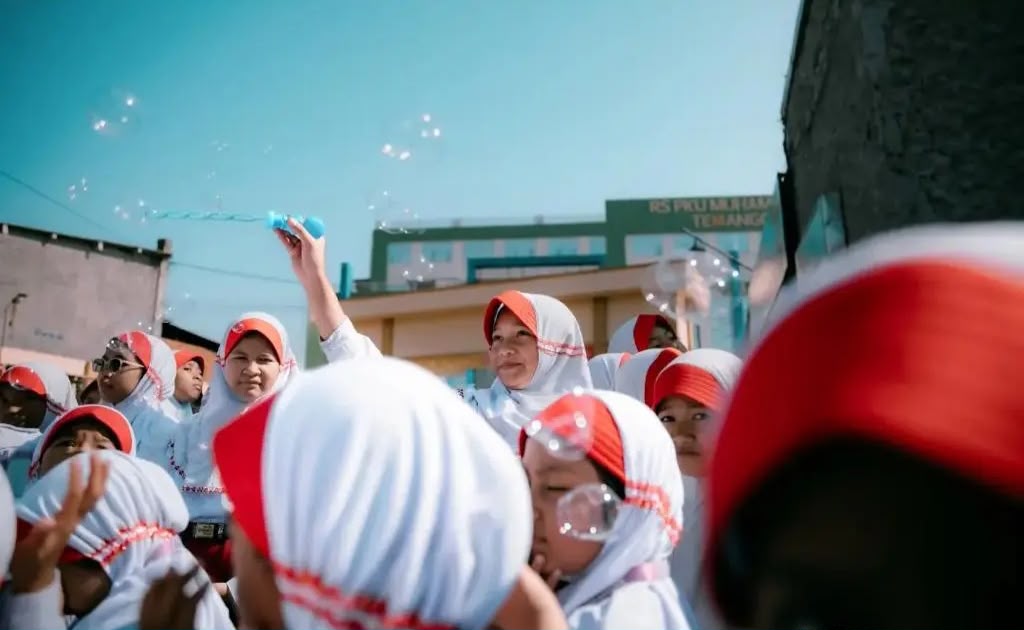 Siswa-siswi SD Muhammadiyah Parakan yang mengenakan seragam pramuka/Hizbul Wathan dan jilbab putih bergaris merah tampak gembira bermain gelembung sabun di bawah langit cerah, mencerminkan lingkungan ramah anak di sekolah favorit temanggung.