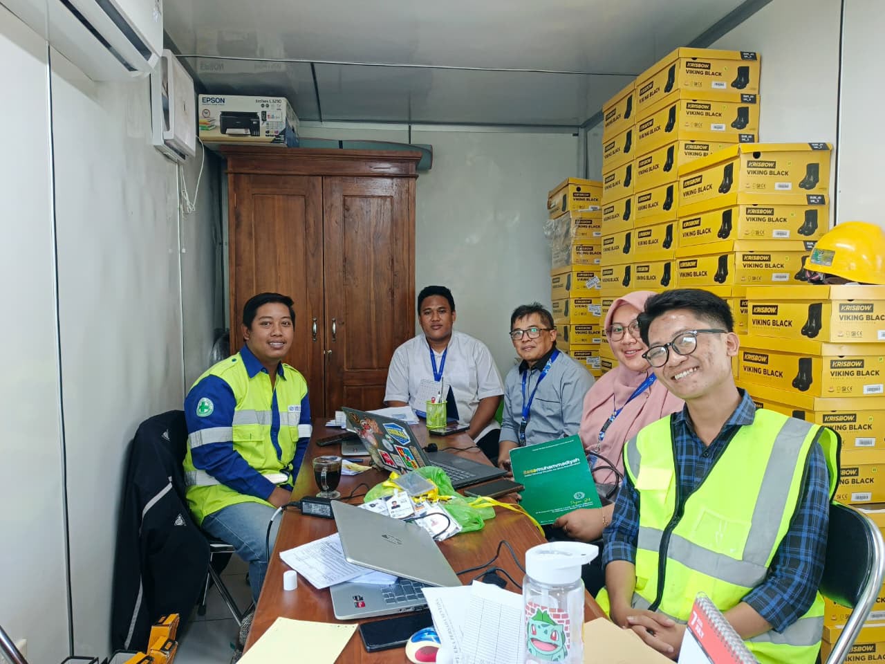 Tim Institut Teknologi Statistika dan Bisnis (ITESA) Muhammadiyah Semarang bertemu perwakilan industri di kantor lapangan di Grobogan. Tumpukan sepatu keselamatan Krisbow dan helm terlihat di latar belakang. Salah satu staf memegang buku berlogo ITESA.