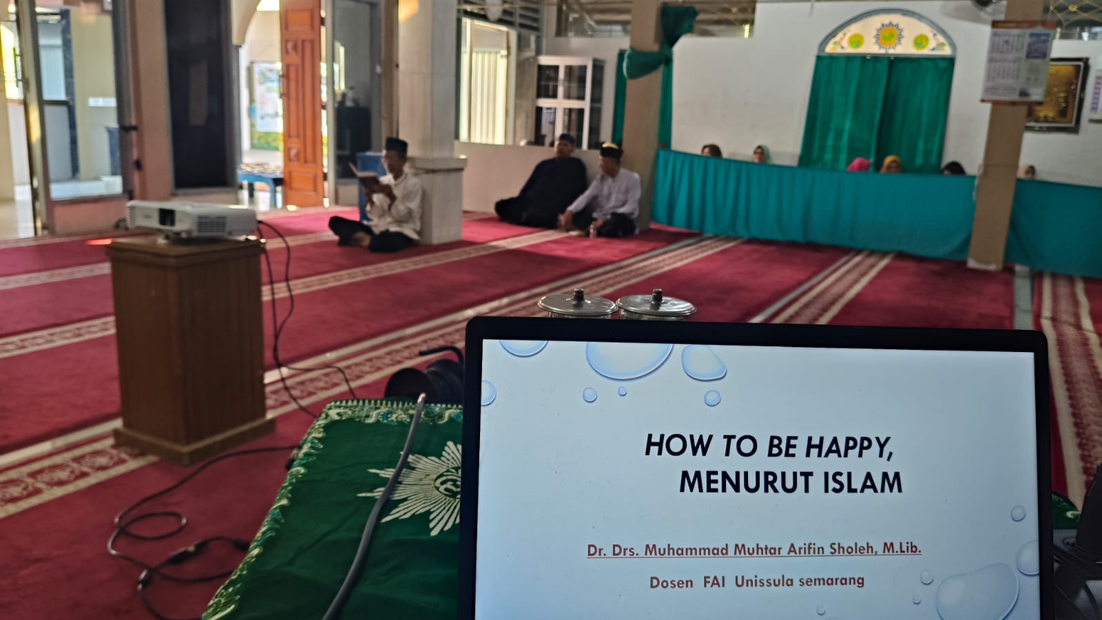 Layar laptop menampilkan slide materi presentasi bertuliskan "HOW TO BE HAPPY, MENURUT ISLAM" oleh Dr. Drs. Muhammad Muhtar Arifin Sholeh, M.Lib. dengan latar suasana ruang shalat Masjid Luqmanul Hakim Tlogosari Kulon Semarang.