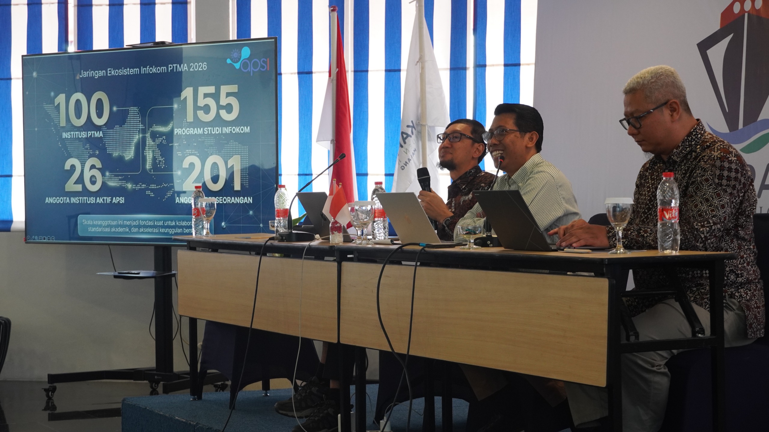 Tiga pimpinan APSI-PTMA duduk di meja panelis dengan latar layar digital yang menampilkan grafik data "Jaringan Ekosistem Infokom PTMA 2026" meliputi 100 institusi dan 155 prodi.
