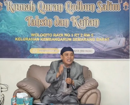 Ustadz Dr. Aang Kunaepi, M.Ag. mengenakan jas abu-abu dan peci hitam sedang memegang mikrofon dan memberikan ceramah di depan spanduk bertuliskan Rumah Quran Qolbun Salim.