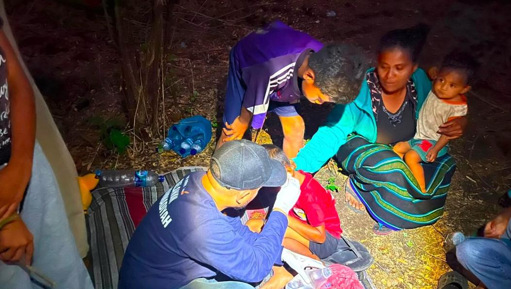 Petugas medis Muhammadiyah mengenakan kaus biru gelap dan sarung tangan medis sedang memeriksa luka pada seorang anak di atas tikar pengungsian pada malam hari, didampingi ibu dan anak-anak lainnya.