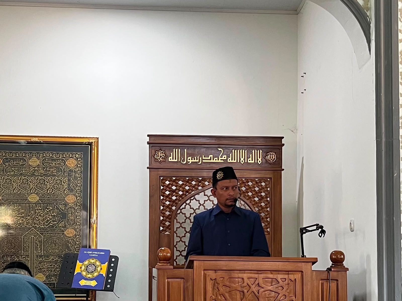 Ustadz Puji, S.Pd.I. mengenakan kemeja biru tua dan peci hitam dengan logo matahari Muhammadiyah berdiri di balik mimbar kayu menyampaikan Khutbah Jumat.