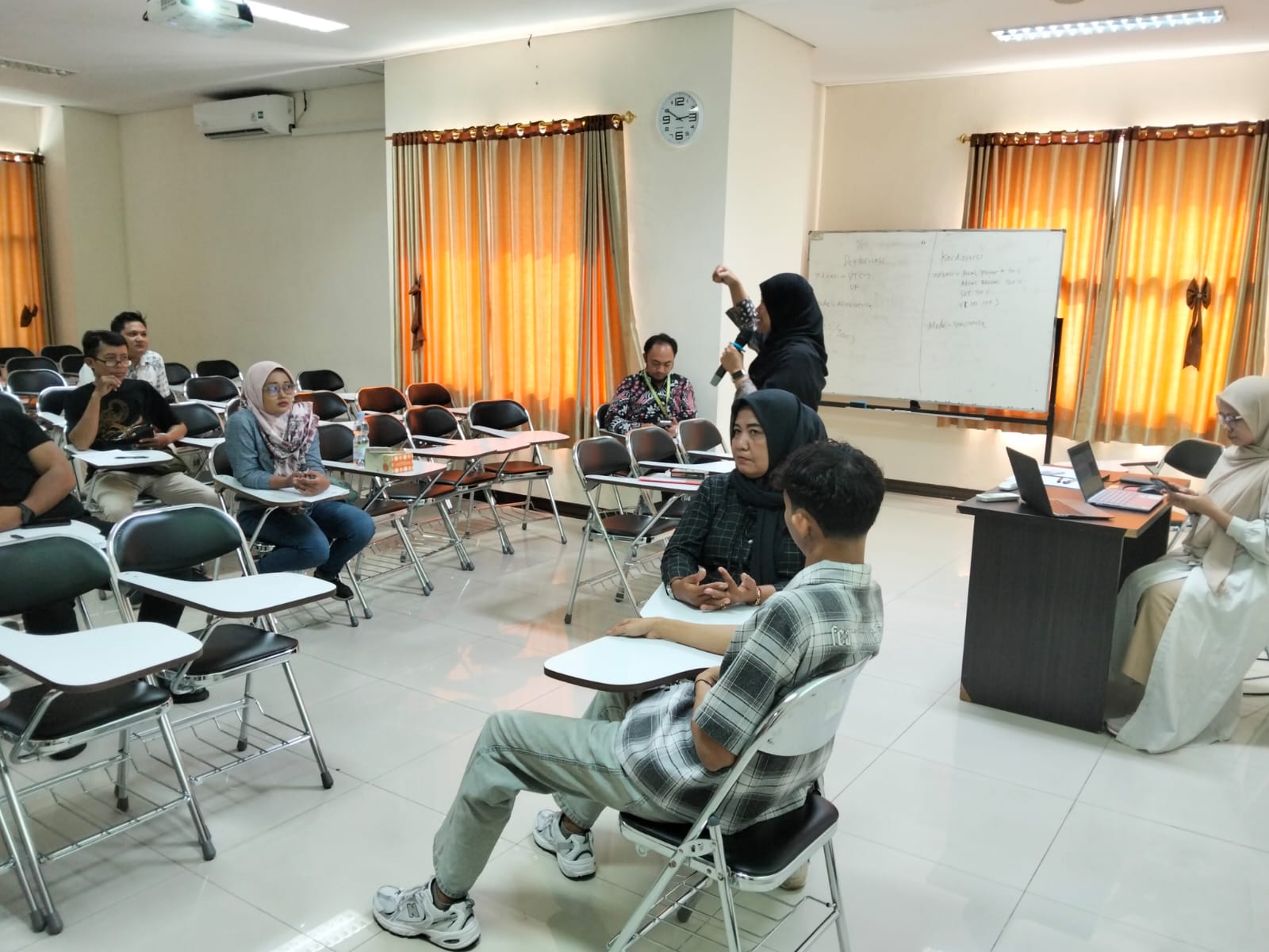 Dosen pakar keperawatan UNIMUS berdiri memegang mikrofon memberikan pengarahan di depan papan tulis, sementara peserta pelatihan dan pemeran pasien bayangan berkumpul di ruang kelas ber-AC mendengarkan instruksi.