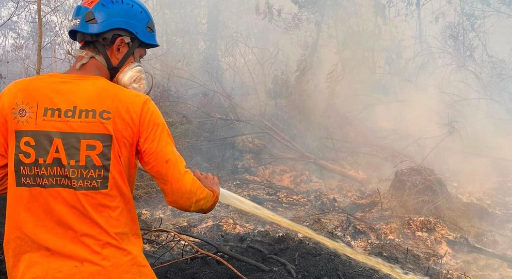 Relawan SAR MDMC Kalbar mengenakan seragam oranye, helm keselamatan biru, dan masker respirator sedang mengarahkan selang air ke semak belukar yang terbakar dan berasap pekat.