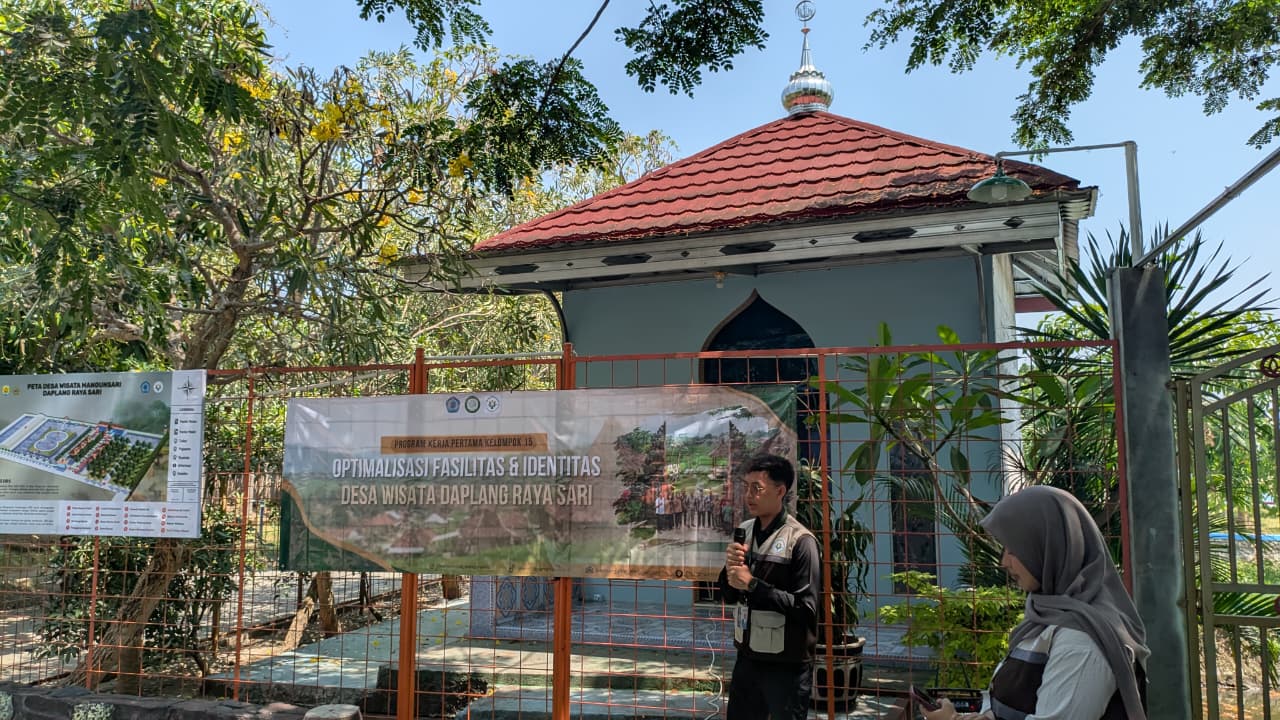 Perwakilan mahasiswa KKN UNIMUS memegang mikrofon menyampaikan sambutan di depan spanduk program kerja dan papan peta navigasi visual kawasan Desa Wisata Daplang Raya Sari.