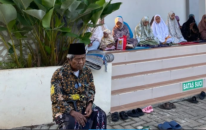 Sosok KH. Salim Abdur Rozaq mengenakan peci hitam dan kemeja batik, duduk memegang tongkat di area halaman Masjid At Taqwa Ngaliyan dengan latar belakang jemaah perempuan.