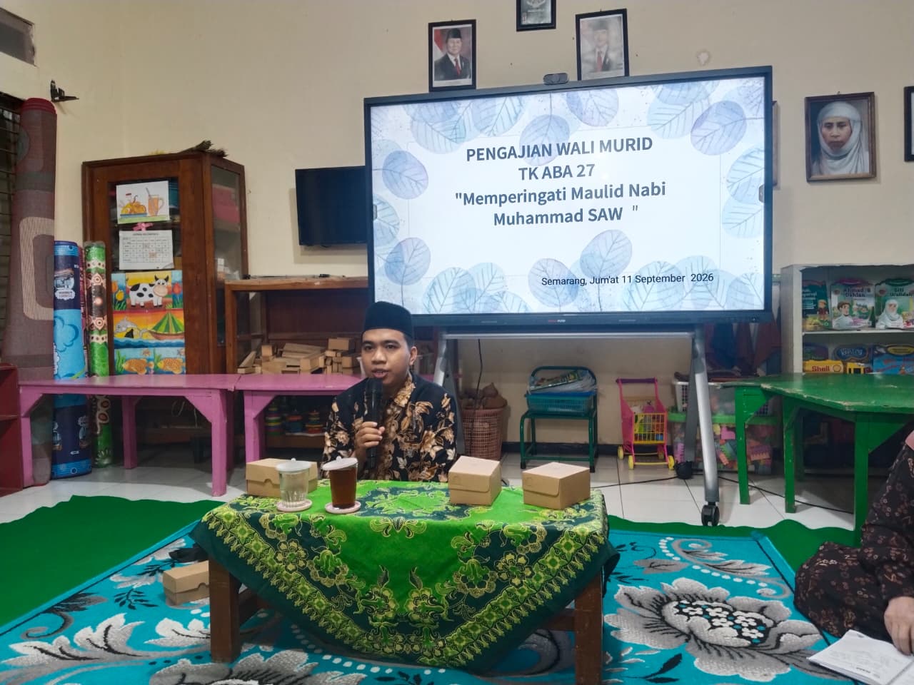 Ust. Muhammad Burhan Khaerudin mengenakan peci hitam dan baju batik sedang membaca materi di hadapan layar presentasi parenting TK 'Aisyiyah Bustanul Athfal.