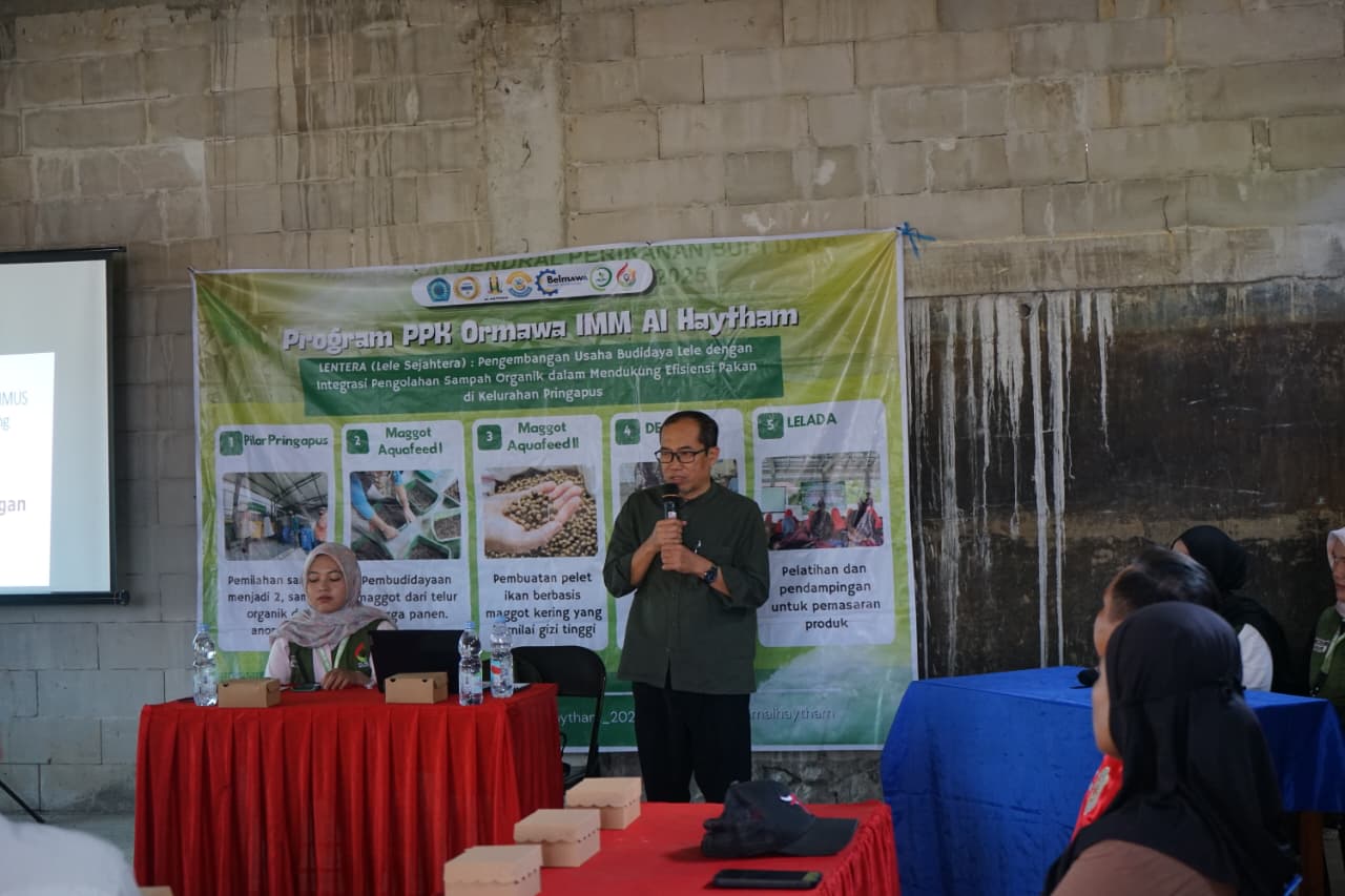 Dr. Agus Suyanto memegang mikrofon menyampaikan materi legalitas halal di depan MMT spanduk bertuliskan Program PPK Ormawa IMM Al Haytham di Aula Kelurahan Pringapus, disamping seorang mahasiswa yang mengoperasikan laptop.