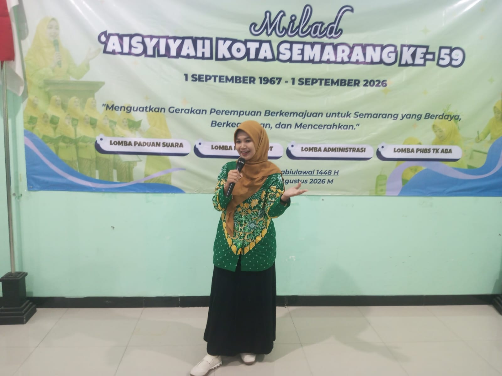 Mahmudah mengenakan batik seragam 'Aisyiyah hijau dan kerudung cokelat berdiri memegang mikrofon dengan ekspresi tersenyum di depan spanduk MMT Milad 'Aisyiyah Kota Semarang ke-59.