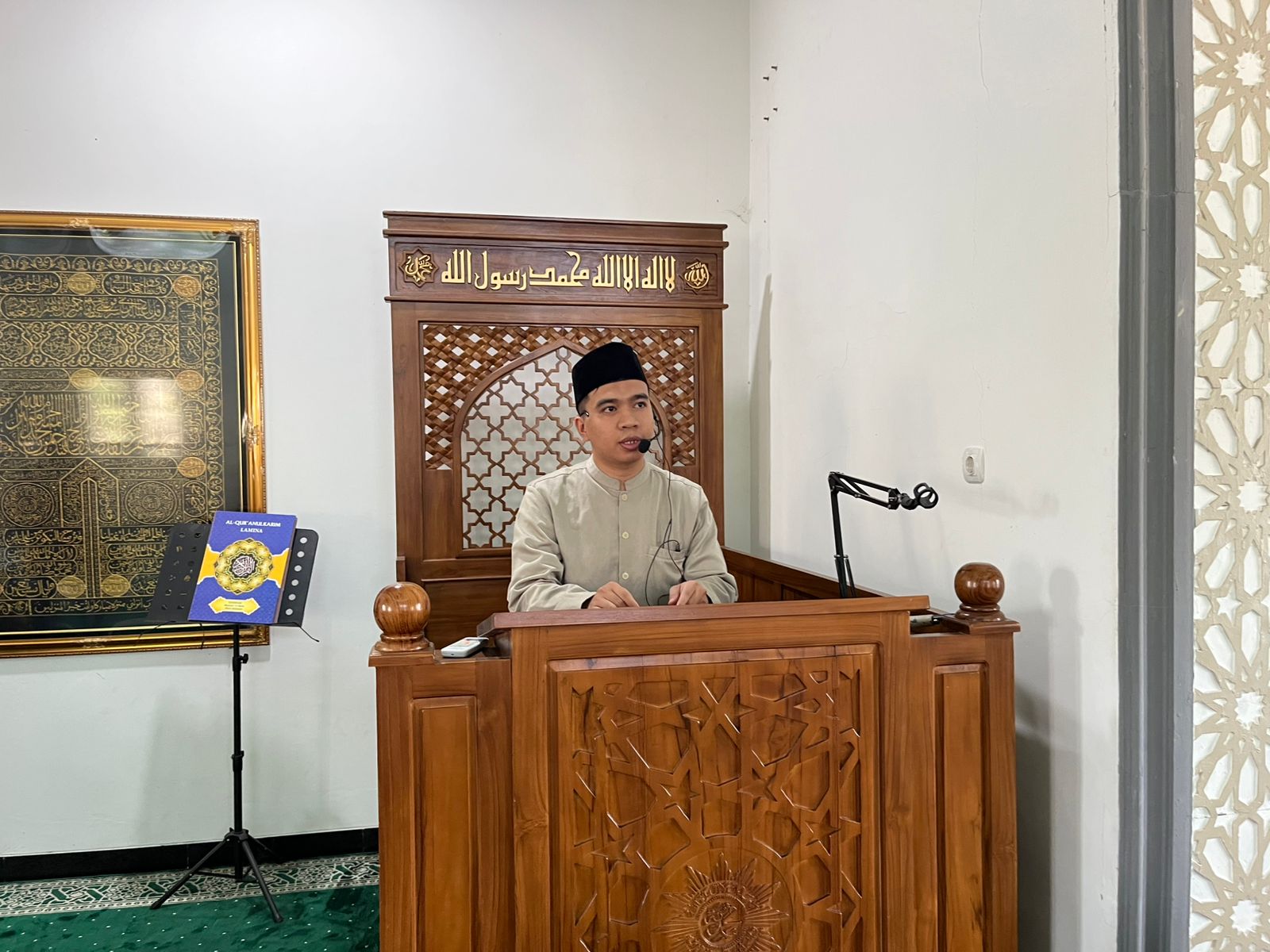 Seorang khatib pria muda mengenakan peci hitam dan baju koko krem sedang menyampaikan khotbah Jumat dari mimbar kayu berukir di dalam Masjid At-Taqwa Wates. Di belakangnya terdapat kaligrafi Islam emas.