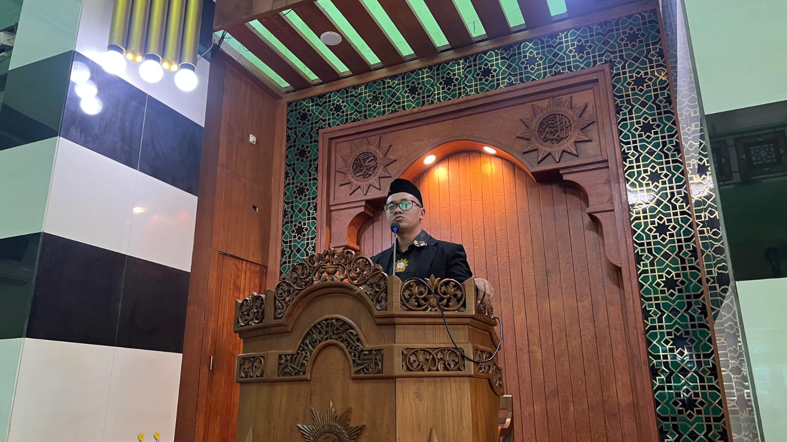 Al Ustadz Raad Noor Fatath mengenakan jas hitam dan peci berdiri di atas mimbar kayu ukir Masjid At-Taqwa PDM Kota Semarang saat menyampaikan khotbah Jumat.