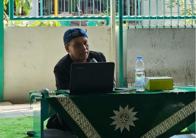 Dr. Aang Kunaepi, M.Ag. mengenakan pakaian hitam dan peci/kupluk biru dengan kacamata di atasnya sedang menyampaikan materi di depan laptop pada meja bertaplak hijau lambang Muhammadiyah.