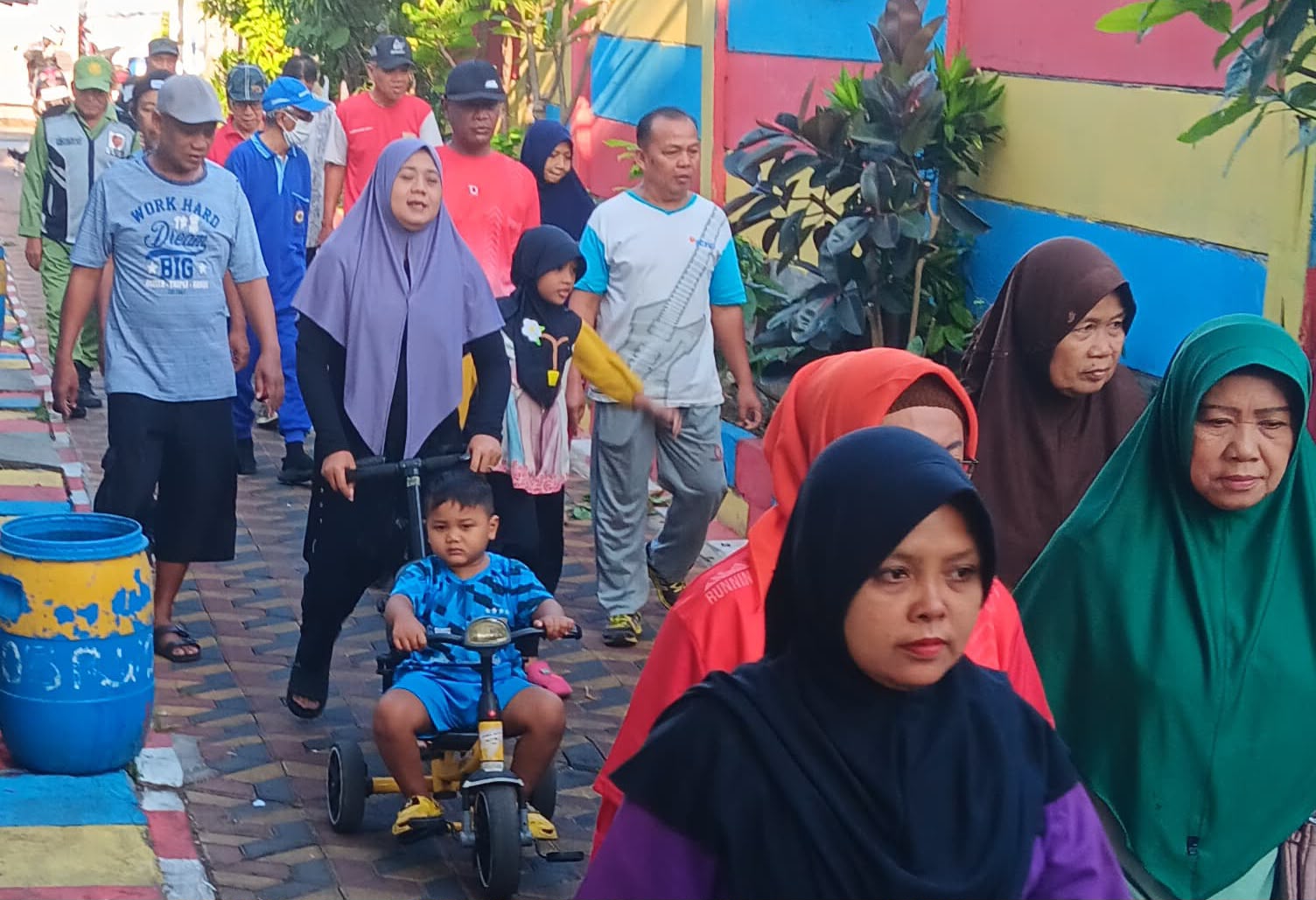 Rombongan warga lintas usia dari anak-anak, pemuda, hingga lansia berjalan santai bersama di gang/jalan permukiman yang penuh warna.