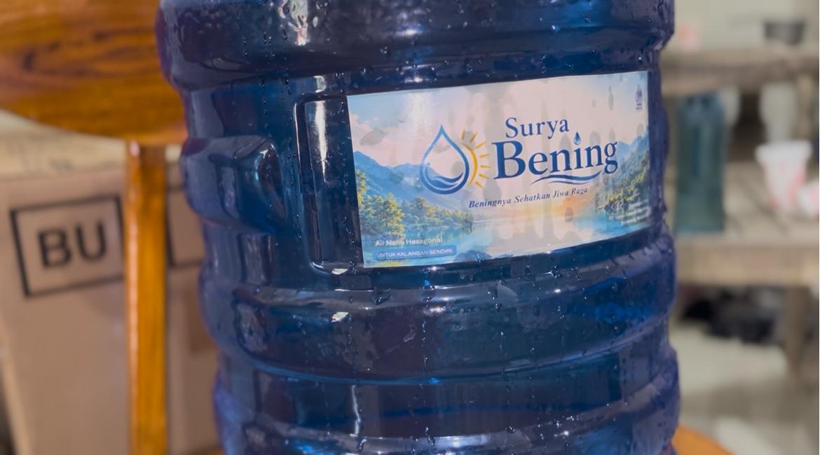 Sebuah galon air minum berwarna biru transparan dengan label merek Surya Bening yang menampilkan gambar tetesan air dan latar pemandangan alam.
