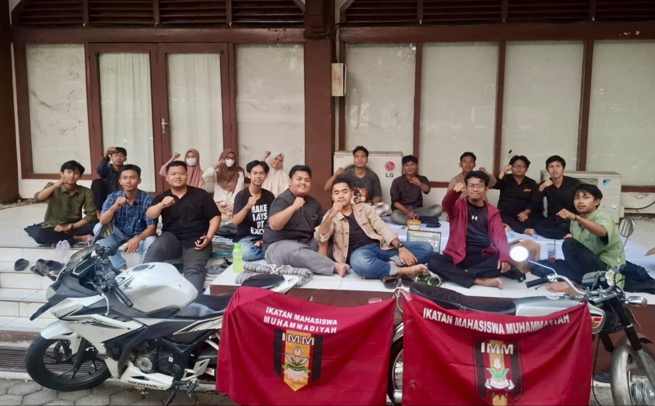 Sembilan belas kader mahasiswa IMM duduk bersama di selasar gedung dengan membentangkan bendera merah Ikatan Mahasiswa Muhammadiyah.
