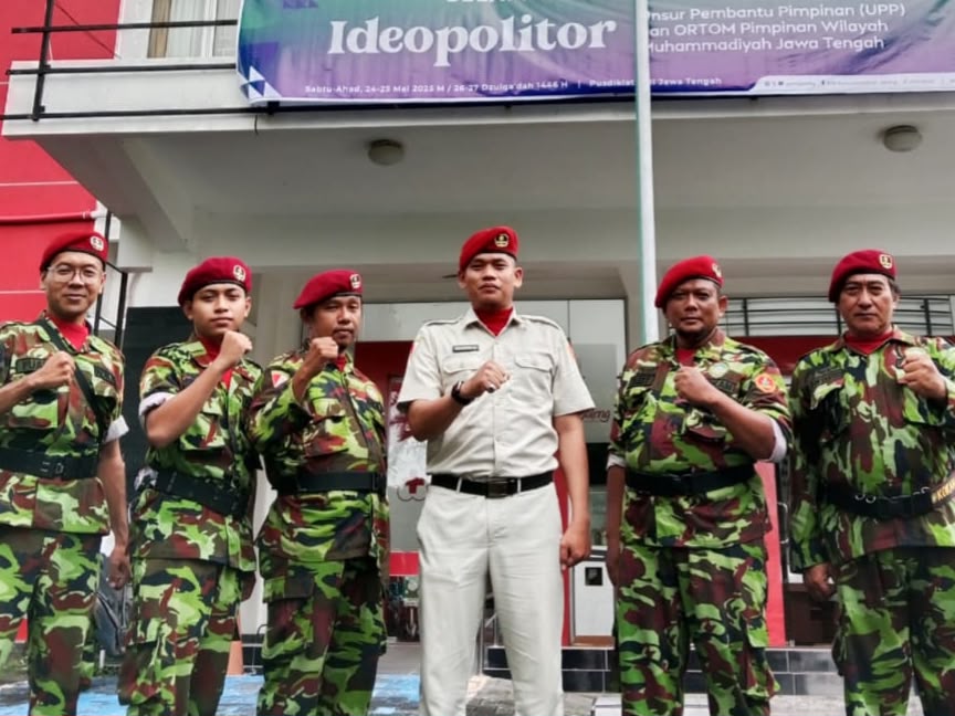 Enam personel KOKAM berbaret merah berdiri sejajar mengepalkan tangan di depan gedung, dengan Komandan KOKAM berdiri di tengah mengenakan seragam PDH berwarna terang.