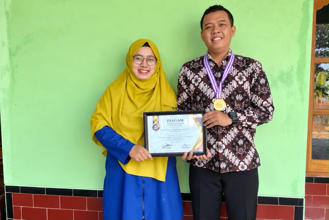 Sepasang akademisi pria dan wanita tersenyum membawa pigura piagam penghargaan dan medali emas dengan latar belakang dinding hijau.