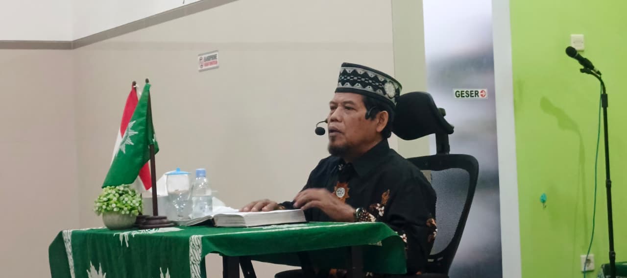 Ustad K.H. Danusiri, M.Ag. mengenakan kemeja batik hitam dan peci bermotif sedang menyampaikan ceramah di meja hijau berlaplak logo Muhammadiyah.