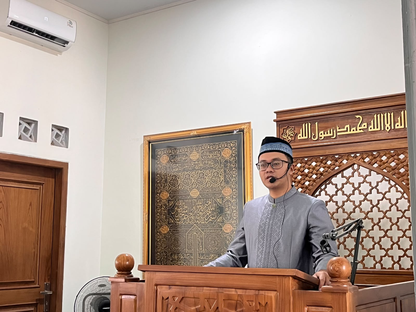 Khatib Ustadz Raad Noor Fattah berkacamata dan mengenakan kemeja koko abu-abu berdiri di atas mimbar kayu Masjid At Taqwa Wates Semarang sambil memegang mikrofon clip-on.