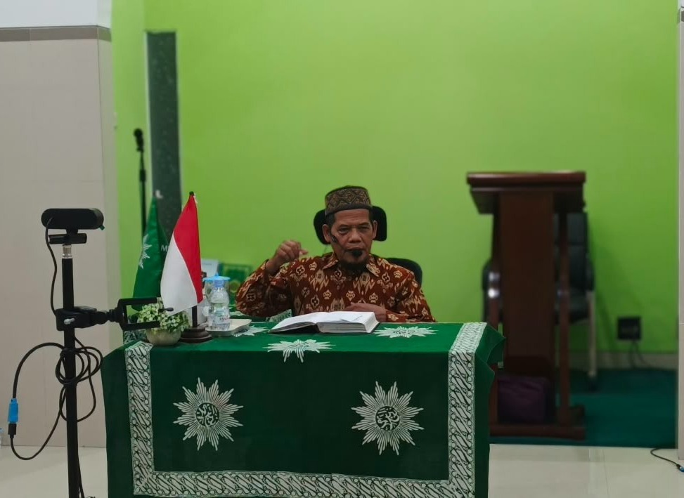 Ustad K.H. Danusiri, M.Ag. berkemeja batik cokelat dan peci motif sedang menyampaikan ceramah dengan gestur tangan di meja berlaplak hijau logo Muhammadiyah.