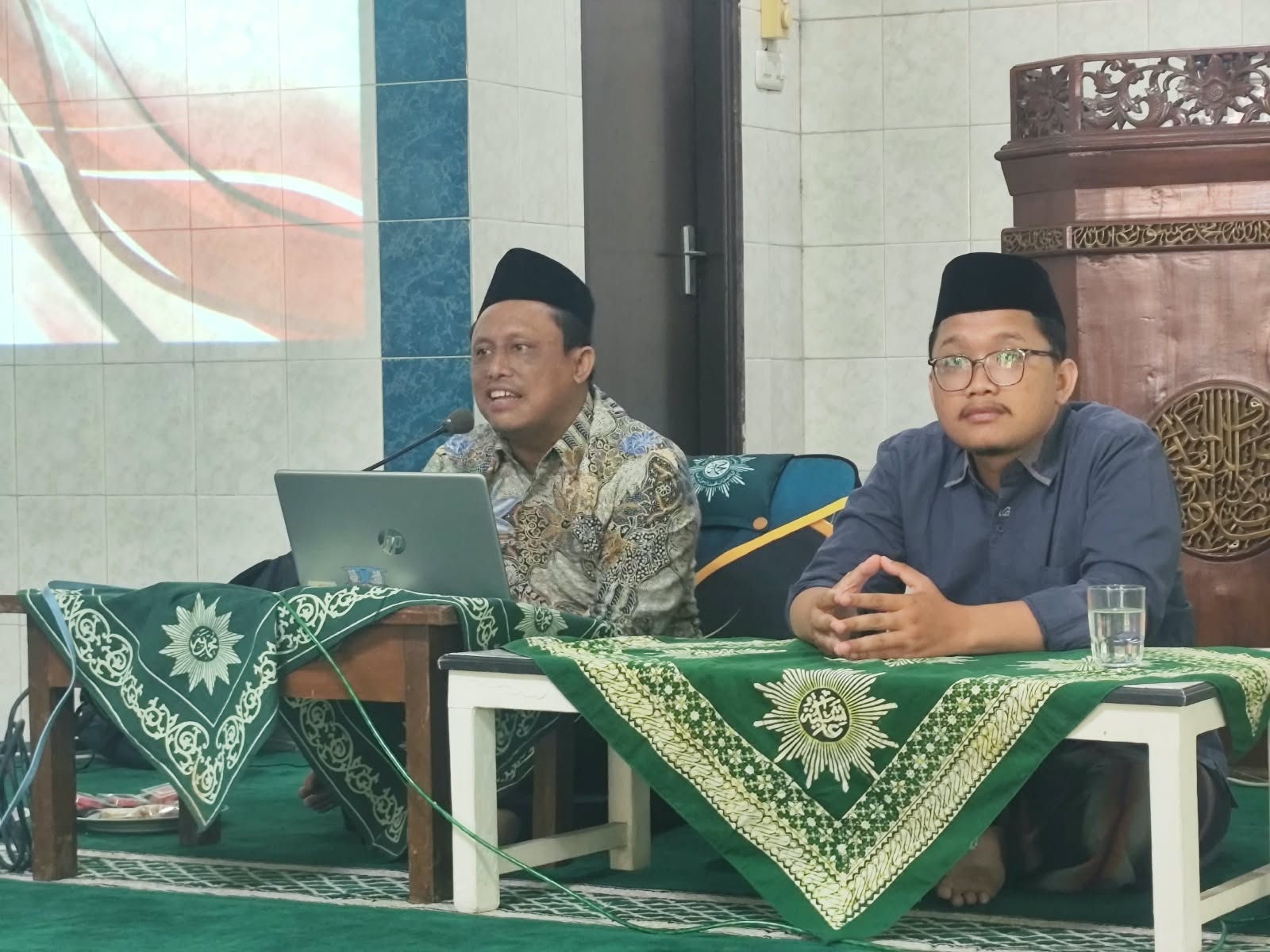 Dua orang narasumber pria berpeci hitam dan berbusana muslim duduk berdampingan di belakang meja berlaplak hijau logo Muhammadiyah di dalam masjid.